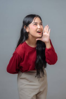 Endonezyalı kadın geniş bir gülümsemeyle elini ağzının yanında tutuyor neşeli ve anlamlı bir jestle. Kırmızı uzun kollu gömlek ve bej pantolon giyiyor. Mutluluk ve enerji saçıyor..