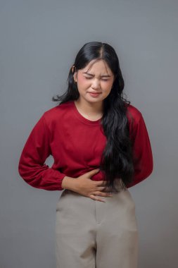 Endonezyalı kadın rahatsızlığını ya da acısını ifade ediyor, tek eliyle karnını tutuyor. Kırmızı uzun kollu gömlek ve bej pantolon giyiyor. Ciddi ve endişeli bir ifade sergiliyor..