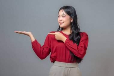 Kırmızı tişörtlü ve bej pantolonlu Asyalı kadın sağ işaret parmağıyla sol eline doğru gülümsüyor ve heyecanını ve coşkusunu ifade ediyor. Tarafsız bir arkaplan ile stüdyo çekimi.