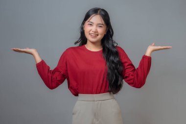 Kırmızı tişörtlü ve bej pantolonlu Asyalı bir kadın, kollarını dışa doğru uzatarak gülümsüyor ve hoş bir jest yapıyor. Tarafsız bir arkaplan ile çekilen bir stüdyoda kendine güvenen, dostane bir ifade..