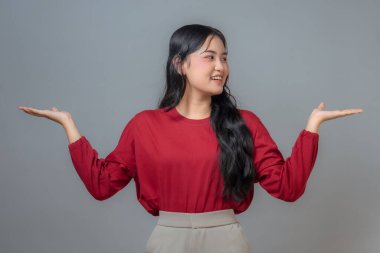 İki eliyle işaret ederken gülümseyen Asyalı bir kadın. Avuçları sanki bir şey sunuyormuş gibi açık. Uzun kollu kırmızı bir bluz ve bej pantolon giyiyor. Gri bir arkaplanda duruyor..