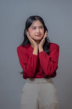 Kırmızı uzun kollu gömlek ve bej pantolon giyen Endonezyalı bir Asyalı kadın. Elleri yanaklarında düşünceli ve ilgili bir ifade sergiliyor. Stüdyo görüntüsü gri arkaplanlı.