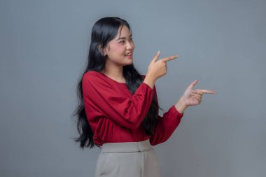 Endonezyalı bir Asyalı kadın kırmızı uzun kollu gömlek ve bej pantolon giyiyor, her iki eli de farklı yönlerde gülümsüyor ve işaret ediyor. Gri arkaplanlı bir stüdyo çekimi, kendinden emin bir hareket..
