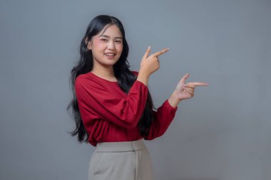 Kırmızı uzun kollu gömlekli ve bej pantolonlu bir Endonezyalı kadın her iki elini de farklı yönlere doğrulturken gülümsüyor. Stüdyo ortamında gri bir geçmişi olan neşeli ve kendinden emin bir jest..