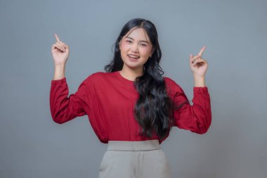 Endonezyalı bir Asyalı kadın gülümsüyor ve iki elini de havaya kaldırarak neşeli bir jest yapıyor. Kırmızı uzun kollu gömlek ve bej pantolon giyiyor. Stüdyo ortamında gri bir arka planı var..