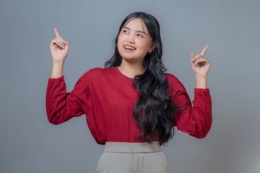 Kırmızı uzun kollu gömlekli, bej pantolonlu, gülümseyen ve iki eliyle yukarı doğru gösteren Endonezyalı bir Asyalı kadın. Tarafsız gri bir arka plana karşı neşeli bir ifadeyle kendinden emin bir şekilde poz verir..