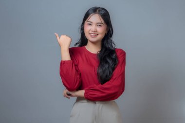 Kırmızı gömlekli ve bej pantolonlu gülümseyen Endonezyalı kadın sağ eliyle baş parmak işareti yapıyor. Gri bir geçmişi olan bir stüdyoda özgüvenli ve pozitif bir vücut dili..