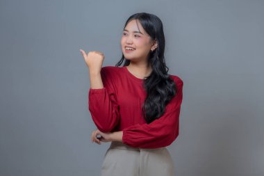 Kırmızı gömlekli ve bej pantolonlu gülümseyen Endonezyalı kadın sağ eliyle baş parmak işareti yapıyor. Gri arkaplanı olan bir stüdyoda özgüvenli ve pozitif bir ifade..