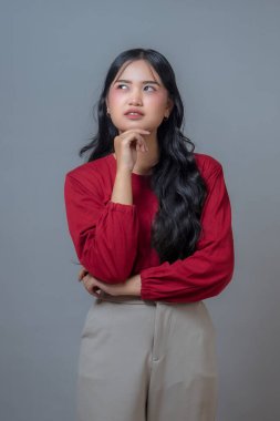 Kırmızı tişörtlü ve bej pantolonlu Endonezyalı kadın, çenesini elinde dinlendirirken düşünceli bir ifadeyle yukarı bakıyor. Gri arkaplanlı stüdyo çekimi, tefekkür ve merak iletiliyor.