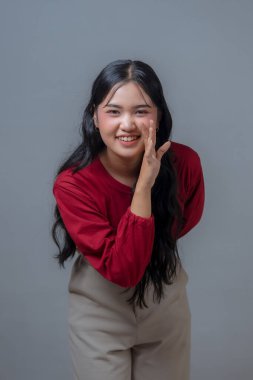 Endonezyalı kadın, eli ağzının yanındayken fısıldayarak gülümserken. Kırmızı bluzlu genç Asyalı kadın gri arka planda şakacı poz veriyor..