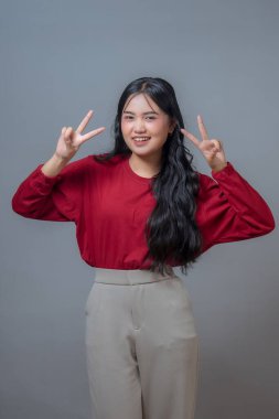 Uzun siyah saçlı, kırmızı bluz ve bej pantolonlu neşeli Asyalı kadın. Gülümsüyor ve iki eliyle birden barış işareti yapıyor. Gri arka planda duruyor..