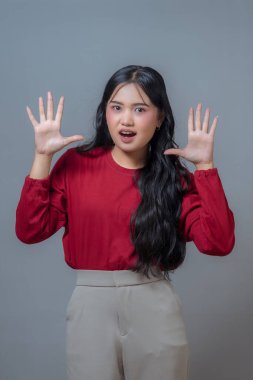 Şaşırmış Endonezyalı genç kadın kırmızı bluz ve bej pantolon giyiyor, iki elini de açık avuç içiyle kaldırıyor, on parmak gösteriyor, ifadesel yüz, düz gri arka planda duruyor..