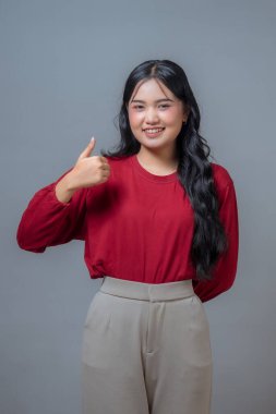 Kırmızı uzun kollu gömlek ve bej pantolon giyen kendine güvenen genç Endonezyalı kadın gülümserken başparmağını kaldırıyor. Gri arka planda stüdyo çekimi, pozitiflik ve onayı ifade eder.