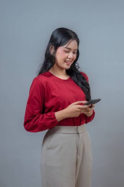 Kırmızı bluzlu ve bej pantolonlu gülümseyen Endonezyalı kadın gri arka planda yan duran akıllı telefon kullanıyor. Rahat duruş ve keyifli görünüş modern dijital yaşam tarzını yakalar..