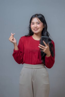 Kırmızı bluz ve bej pantolon giymiş gülümseyen Endonezyalı bir kadın bir elinde akıllı bir telefon tutarken Koreli bir parmak kalp hareketi yapıyor, dijital bağlamda sevgi ve dostluk gösteriyor..