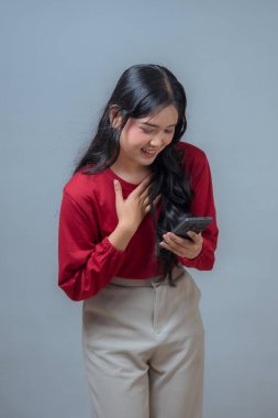 Kırmızı bluz ve bej pantolon giyen Endonezyalı bir kadın akıllı telefonuna bakarken sıcak bir şekilde gülümser, bir elini göğsüne koyar neşe, minnettarlık ya da duygusal bir tepki olarak.