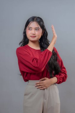 Kırmızı bluzlu ve bej pantolonlu Endonezyalı genç bir kadın, elini kaldırarak, dostça bir gülümsemeyle selamlıyor ya da ilgi gösteriyordu. Gri bir arka planda izole edilmiş, kendine güvenini gösteriyor..