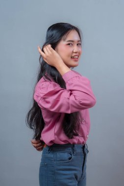 Gülümseyen Asyalı genç bir Endonezyalı kadının doğal olarak saçlarına dokunuşunu, pembe bluz ve kot pantolon giyişini, sıradan, gri bir arka planda duruşunu..