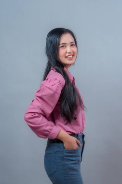 Minimalist bir stüdyoda elleri ceplerinde pembe bir bluz ve kot pantolon giymiş, kameraya içten gülümseyen Asyalı genç bir kadının yan portresi..