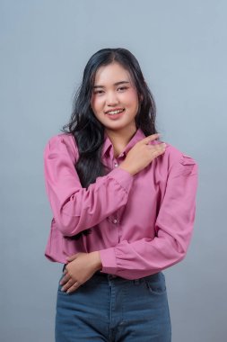 Uzun siyah saçlı, pembe gömlekli ve mavi kotlu gülümseyen Endonezyalı genç bir kadının portresi, tarafsız bir arkaplana karşı omuz omuza poz veriyor..