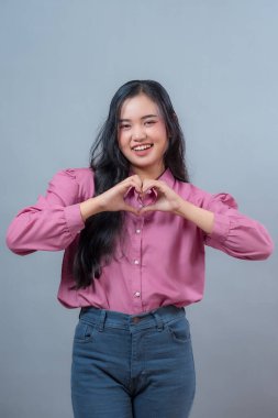 Uzun siyah saçlı, güzel, gülümseyen Endonezyalı kadın elleriyle kalp hareketi yapıyor. Gri arka planda pembe bir bluz ve kot pantolon giyiyor..