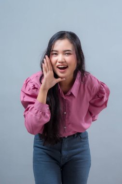 Gülümseyen Endonezyalı kadın pembe gömlekli ve kot pantolonlu sanki arıyor ya da bağırıyor. Stüdyoda gri arka planda poz veren etkileyici genç Asyalı kadın model..