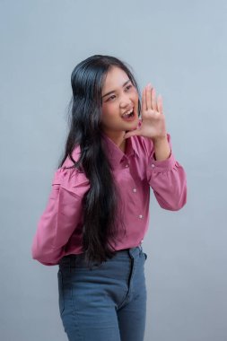 Pembe bluzlu ve kot pantolonlu kendine güvenen genç Endonezyalı kadın elini ağzının yanına koyup bağırıyor. Açık ve sade arkaplanı olan bir stüdyo portresi.