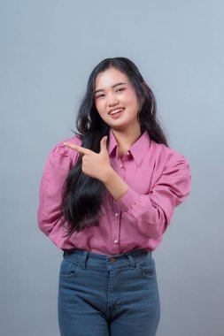 Pembe bluz ve kot pantolon giymiş neşeli bir Endonezyalı kadının portresi. Gri arka planda izole edilmiş, baş parmağıyla kenarı gösterirken güvenle gülümsüyor..