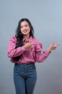 Pembe bluz ve kot pantolon giymiş Endonezyalı genç bir kadının portresi, iki eliyle gülümseyen ve bir tarafı işaret eden, heyecanı ifade eden ve gri arka plana karşı bir şeyleri destekleyen.