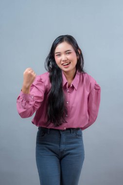 Gülümseyen Endonezyalı kadın pembe bluz ve kot pantolon giyerek neşeli bir jestle yumruğunu kaldırıyor, başarısını, motivasyonunu, coşkusunu ve bir stüdyo ortamında kendine güvenini gösteriyor..