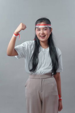 Uzun siyah saçlı Endonezyalı genç bir kadın zafer jesti olarak yumruğunu kaldırırken kendinden emin bir şekilde gülümsüyor. Kırmızı-beyaz saç bandı ve bileklik takarak güç yayıyor..