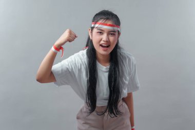 Uzun siyah saçlı, beyaz gömlekli, kırmızı-beyaz bantlı enerjik genç Endonezyalı kadın, öne eğilip gülümsüyor ve gururla yumruğunu kaldırıyor..