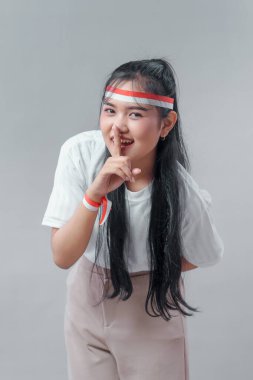 Endonezyalı genç bir kadın kırmızı ve beyaz bir saç bandı ve kurdele takıyor, gülümsüyor ve dudaklarında parmak ile şşş hareketi yapıyor. Oyuncu, kendinden emin, anlamlı, kişilik dolu..