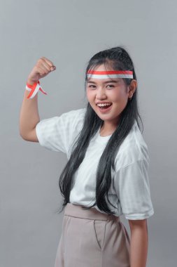 Uzun siyah saçlı neşeli Endonezyalı kadın parlak bir şekilde gülümsüyor ve esnek bir kol pozu veriyor. Beyaz gömlek ve kırmızı-beyaz saç bandı takıyor, ulusal gurur ve enerjiyi temsil ediyor..