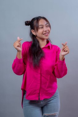 Uzay çörekleri olan Endonezyalı genç bir kadın canlı pembe bir gömlek ve yırtık bir kot pantolon giyerek stüdyoda iki eliyle Kore kalbi hareketi yaparken neşeli bir şekilde gülümsüyor..