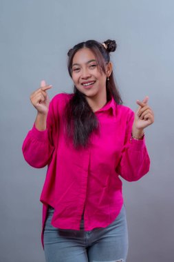 Uzun siyah saçlı, çift katlı saç stili olan parlak pembe bir gömlek ve kot pantolon giyen mutlu Endonezyalı kadın iki eliyle kalp hareketi yapıyor ve neşeyle gülümsüyor..