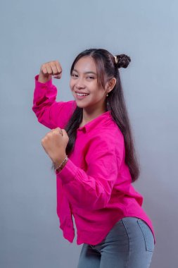 Kollarını esneterek gücünü gösteren mutlu, genç Endonezyalı bir kadının portresi, stüdyo ortamında parlak pembe bir gömlek ve kot pantolon giyerek güvenle gülümsüyor..