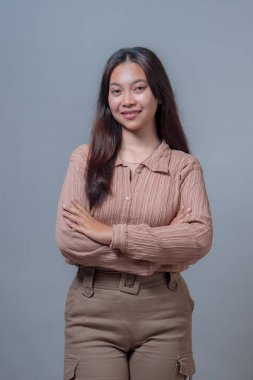 Uzun saçlı, kollarını kavuştururken gülümseyen kendinden emin Endonezyalı bir kadın. Kahverengi desenli bluz ve kargo pantolonu giyiyor. Rahatlamış ama iddialı bir vücut dili gösteriyor..