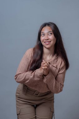 Endonezyalı genç bir Asyalı kadın, elleri birbirine kenetlenmiş, neşeli ve ilginç bir ifadeyle, tarafsız gri bir arkaplana karşı durarak, neşeyle gülümsüyor..