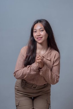 Endonezyalı genç bir kadının portresi. Uzun saçları yumuşak bir şekilde gülümsüyor, elleri sımsıkı bağlı hafifçe öne eğiliyor. Bej kıyafetler giyiyor, sıcaklığını, dostluğunu ve samimiyetini ifade ediyor..