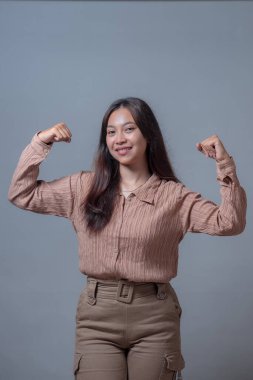 Endonezyalı bir kadın, bej bir bluz ve bej pantolon içinde gülümseyen ve mutlu görünen bir güç gösterisiyle kollarını rahatça esnetiyor..