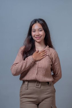 Endonezyalı genç bir kadın içten bir ifadeyle gülümseyerek ellerini göğsüne koyuyor. Minnettarlık ya da samimiyet göstergesi olarak. Gri bir arkaplanda, günlük kıyafetlerle dikiliyor..