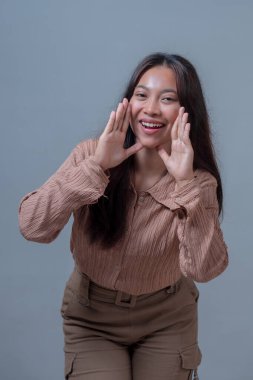 Gülümseyen, bej gömlekli Endonezyalı genç bir kadın, ellerini ağzının yanında şakacı bir şekilde tutuyor, sanki anons ediyor ya da sesleniyor gibi, düz gri bir arka planda duruyor..