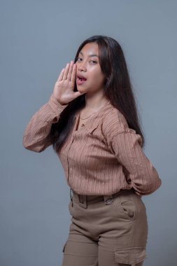 Bej tişörtlü genç Endonezyalı bir kadın, bir eli ağzının yanında duruyor ve bir şey söylüyor ya da anons ediyor gibi hareket ediyor, anlamlı ve kendinden emin bir yüz ifadesiyle..