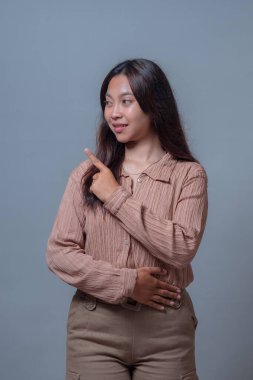 Bej bluzlu Endonezyalı bir kadın bir eli belinde dururken gülümseyip yukarı bakıyor. Gri bir geçmişi olan bir stüdyoda, özgüvenli ve dostane bir tavır sergiliyor..