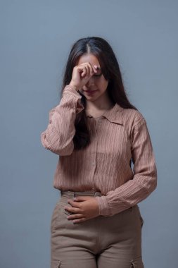 Düşünceli bir Endonezyalı kadın bej bluzlu ve bej pantolonlu, nazikçe alnına dokunuyor, bir anlık yansıma gösteriyor. Gri bir arka planda izole edilmiş, sakin ve içe dönük.