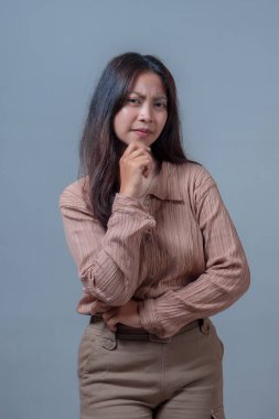 Bej bluzlu ve bej pantolonlu Endonezyalı kadın eli çenesinde düşünceli bir poz veriyor. Gri bir arka planda izole edilmiş, özgüven ve düşünce yayıyor..