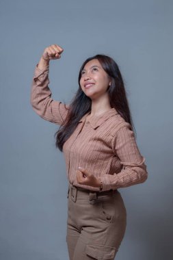 Kendinden emin Endonezyalı genç bir kadın güçlü bir duruşta gülümseyip kollarını esnetiyor. Güç ve iyimserlik gösteriyor. Tarafsız arka planlı stüdyo portresi, gündelik kıyafet ve yukarı bakış..