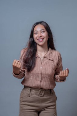 Endonezyalı neşeli genç bir kadının portresi çift yumruk hareketi gösterirken kendine güvenerek gülümsüyor. Mutluluk, motivasyon ve başarıyı ifade eden gri arkaplan üzerine stüdyo çekimi.