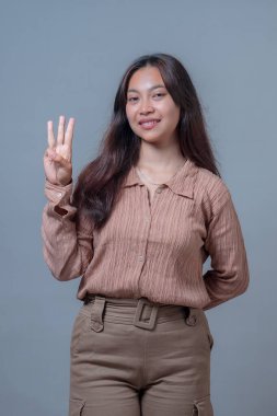 Uzun siyah saçlı Endonezyalı bir kadın üç parmak hareketi yaparken nazikçe gülümsüyor. O, dokulu kahverengi bir gömlek giyer ve düz gri bir arka planda güvenle poz verir..
